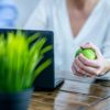 Woman Squeezing A Green Stress Ball Sitting Down At A Desk Next To A Green Plant.