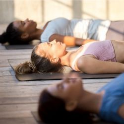Three people lying face upwards on yoga mats against a wooden floor